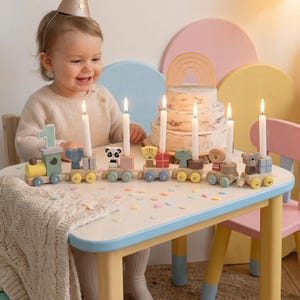 May include: A wooden birthday train with animal-themed candle holders. The train cars feature a number 1, an elephant, a panda, a giraffe, a lion, and a bear. Each car holds a lit white candle. The table is decorated with confetti.