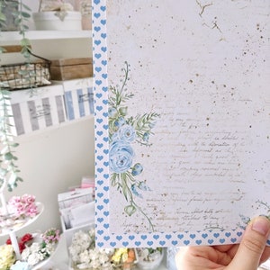 May include: A white card with a blue heart pattern border. The card has a watercolor floral design with blue flowers and green leaves. The card has a vintage look with a faded background and handwritten text. The text reads "They need to be happy, serve the very service of the heart, for it is the devotion of the soul. Deformed as they are, they are so precious, so necessary, so good for taking good care of, and the place reserved for them in the world is remarkable as is the love they command in human hearts. Inspired from the pages of "The Secret Garden" by Frances Hodgson Burnett.  "The roses sprout fully, splendidly, and their fragrance fills the air.  The leaves are attached to long, velvety stems, and they are a wonderful sight to behold."