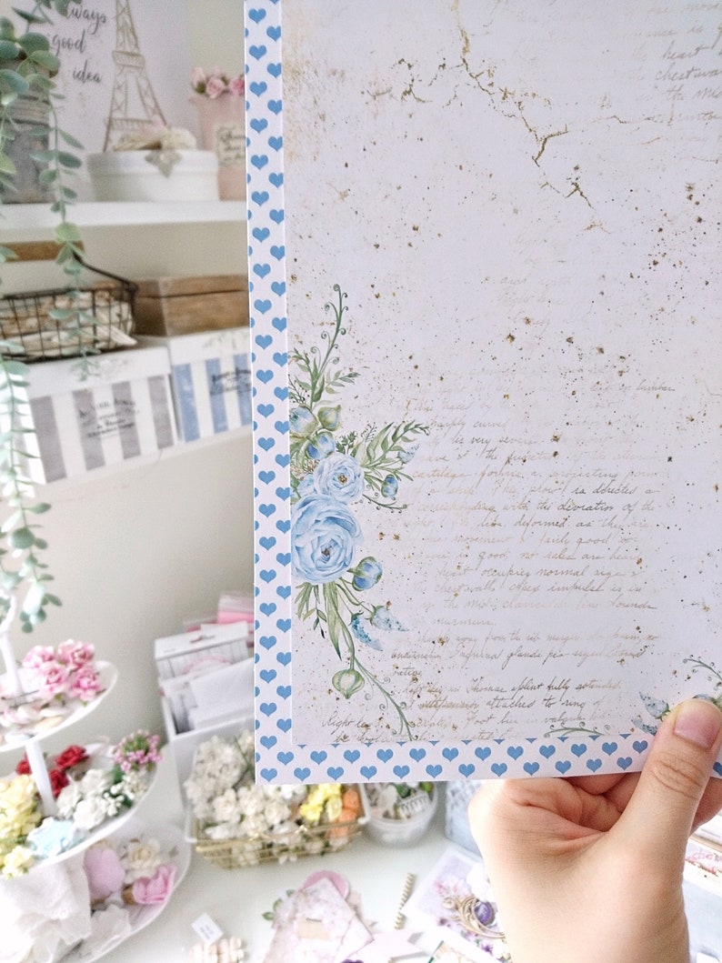 May include: A white card with a blue heart pattern border. The card has a watercolor floral design with blue flowers and green leaves. The card has a vintage look with a faded background and handwritten text. The text reads "They need to be happy, serve the very service of the heart, for it is the devotion of the soul. Deformed as they are, they are so precious, so necessary, so good for taking good care of, and the place reserved for them in the world is remarkable as is the love they command in human hearts. Inspired from the pages of "The Secret Garden" by Frances Hodgson Burnett.  "The roses sprout fully, splendidly, and their fragrance fills the air.  The leaves are attached to long, velvety stems, and they are a wonderful sight to behold."