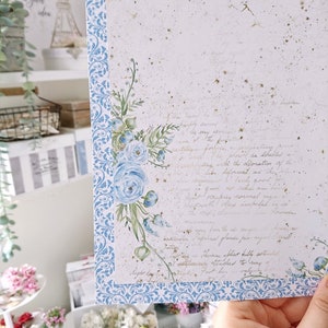 May include: A white card with a blue and white patterned border. The card has a watercolor illustration of blue flowers and leaves in the upper right corner. The card has handwritten text in gold ink.
