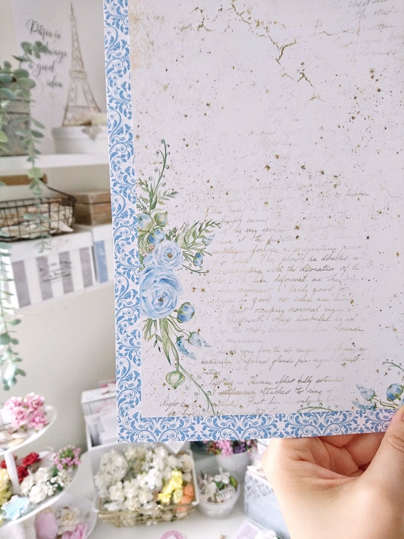 May include: A white card with a blue and white patterned border. The card has a watercolor illustration of blue flowers and leaves in the upper right corner. The card has handwritten text in gold ink.