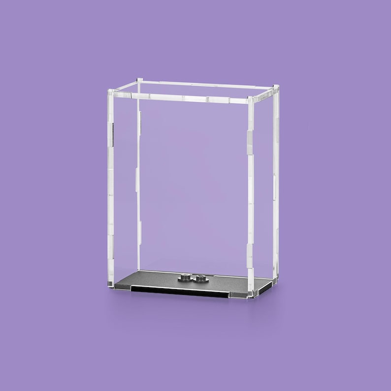 May include: A clear acrylic display case with a rectangular shape. The case has a black base and a clear top frame. The background is a solid purple color. The case is designed to showcase collectibles or other items.