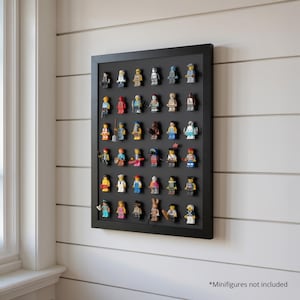 Puede incluir: Un expositor enmarcado en negro con un fondo negro, que muestra una colección de coloridas minifiguras. El expositor está montado en una pared de paneles blancos. El texto en la parte inferior dice "*Minifiguras no incluidas."
