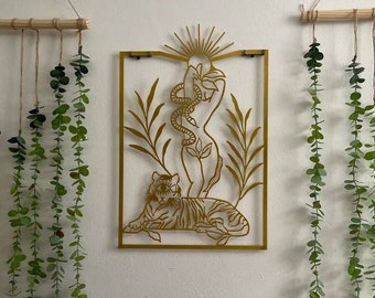 May include: Gold metal wall art featuring a stylized image of a woman with a snake wrapped around her, a tiger, and floral elements. The art is framed with a thin metal border.
