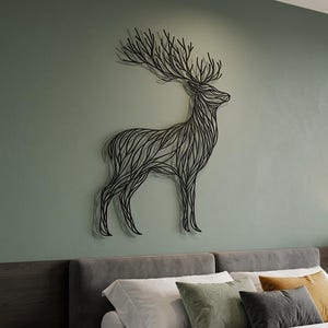 May include: A black metal deer wall art decorates a green wall above a bed with gray headboard and pillows. The deer is stylized with a line art design. The bed has white sheets and a brown blanket.