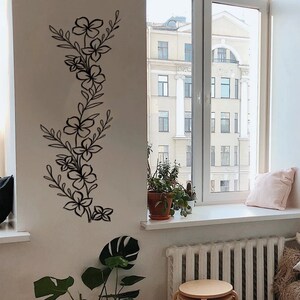 Puede incluir: Un interior de sala de estar con una pared blanca que presenta una calcoman&iacute;a de pared floral negra. Una ventana con vista a un edificio de la ciudad est&aacute; en el lado derecho de la imagen. Un sof&aacute; con una manta de tiro marr&oacute;n peluda est&aacute; en primer plano. Una guitarra est&aacute; apoyada contra el sof&aacute;.