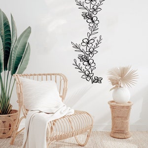 Puede incluir: Una silla de mimbre blanca con una manta blanca y una almohada blanca se encuentra frente a una pared blanca con una calcoman&iacute;a floral negra. Una mesa auxiliar de mimbre con un jarr&oacute;n blanco y una palmera seca se encuentra a la derecha de la silla.