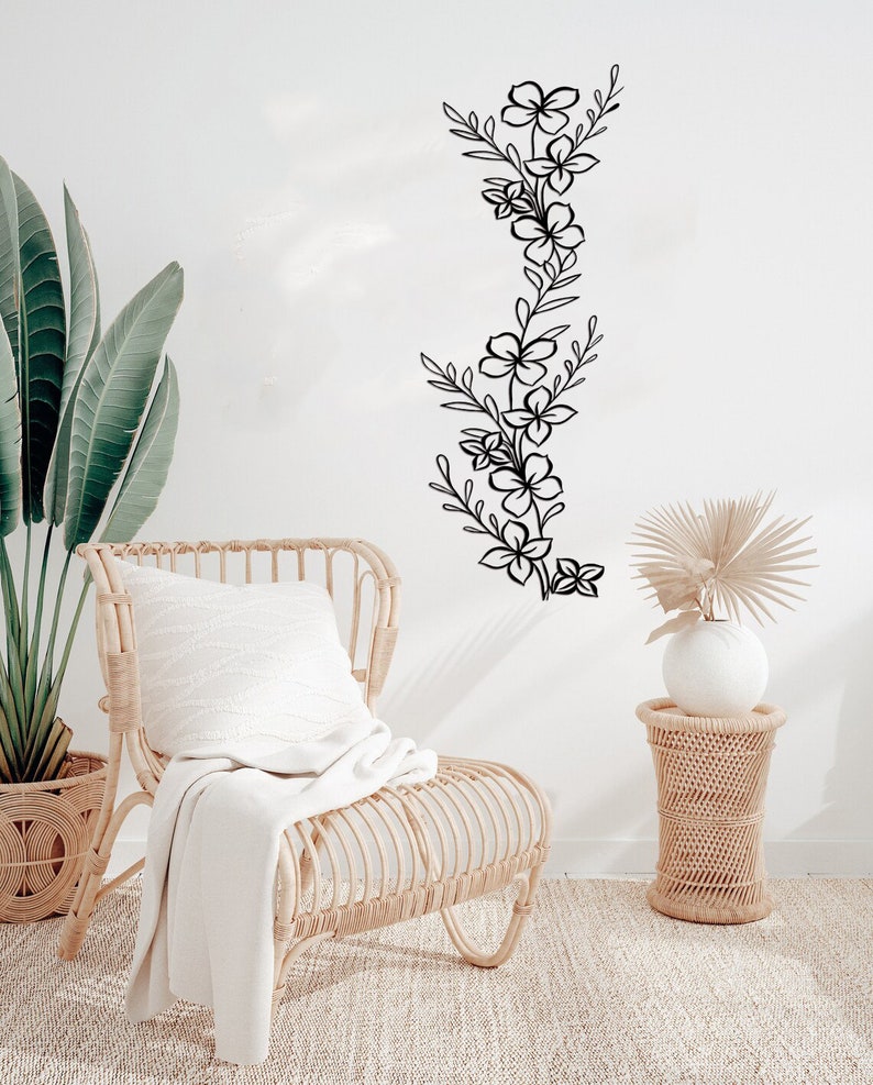 Puede incluir: Una silla de mimbre blanca con una manta blanca y una almohada blanca se encuentra frente a una pared blanca con una calcoman&iacute;a floral negra. Una mesa auxiliar de mimbre con un jarr&oacute;n blanco y una palmera seca se encuentra a la derecha de la silla.