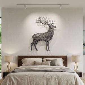 May include: A black metal deer wall art decorates a bedroom wall above a bed with neutral-toned bedding. The deer sculpture has detailed lines and large antlers. Two lamps with gold bases sit on either side of the bed.