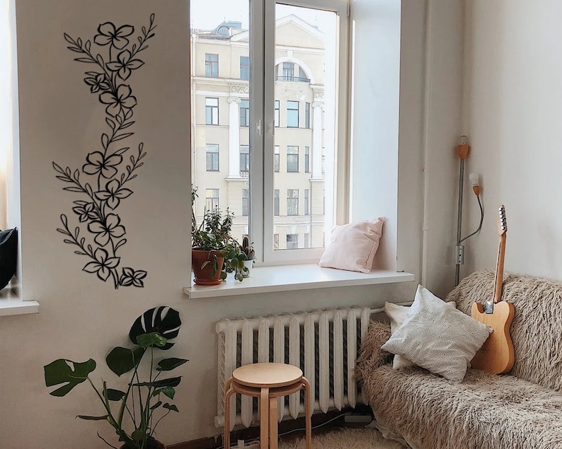Puede incluir: Un interior de sala de estar con una pared blanca que presenta una calcoman&iacute;a de pared floral negra. Una ventana con vista a un edificio de la ciudad est&aacute; en el lado derecho de la imagen. Un sof&aacute; con una manta de tiro marr&oacute;n peluda est&aacute; en primer plano. Una guitarra est&aacute; apoyada contra el sof&aacute;.