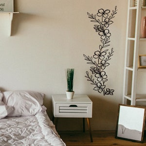 Puede incluir: Una pared blanca con una calcoman&iacute;a de pared floral negra. La calcoman&iacute;a presenta una disposici&oacute;n vertical de flores y hojas estilizadas. Un estante blanco con el texto "And see what happens" est&aacute; por encima de la calcoman&iacute;a. Una mesita de noche blanca con una peque&ntilde;a planta en maceta est&aacute; en primer plano.