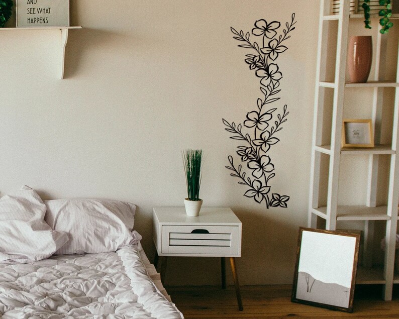 Puede incluir: Una pared blanca con una calcoman&iacute;a de pared floral negra. La calcoman&iacute;a presenta una disposici&oacute;n vertical de flores y hojas estilizadas. Un estante blanco con el texto "And see what happens" est&aacute; por encima de la calcoman&iacute;a. Una mesita de noche blanca con una peque&ntilde;a planta en maceta est&aacute; en primer plano.
