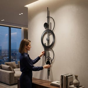 May include: A black metal wall art piece with a geometric design of circles, lines, and semi-circles. The artwork is mounted on a light-coloured wall. A woman is seen adjusting the artwork.