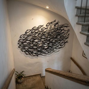 May include: A large black metal wall art piece featuring a stylized school of fish swimming in a dense, flowing formation. The design consists of numerous interconnected fish silhouettes with thin, curved lines creating a sense of movement.