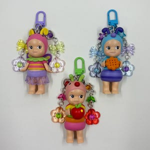May include: Three colorful keychains featuring small doll figures. Each doll has a unique outfit and accessories, including flower and butterfly accents. The keychains have a metal clasp for easy attachment to bags or keys.