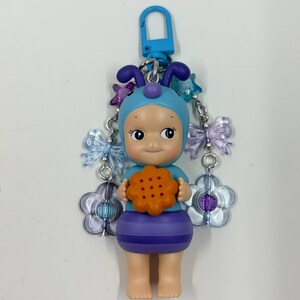 May include: A blue and purple keychain featuring a doll-like figure wearing a blue bonnet with antennae and holding an orange cookie. The keychain has a blue clasp and is adorned with flower and butterfly charms.