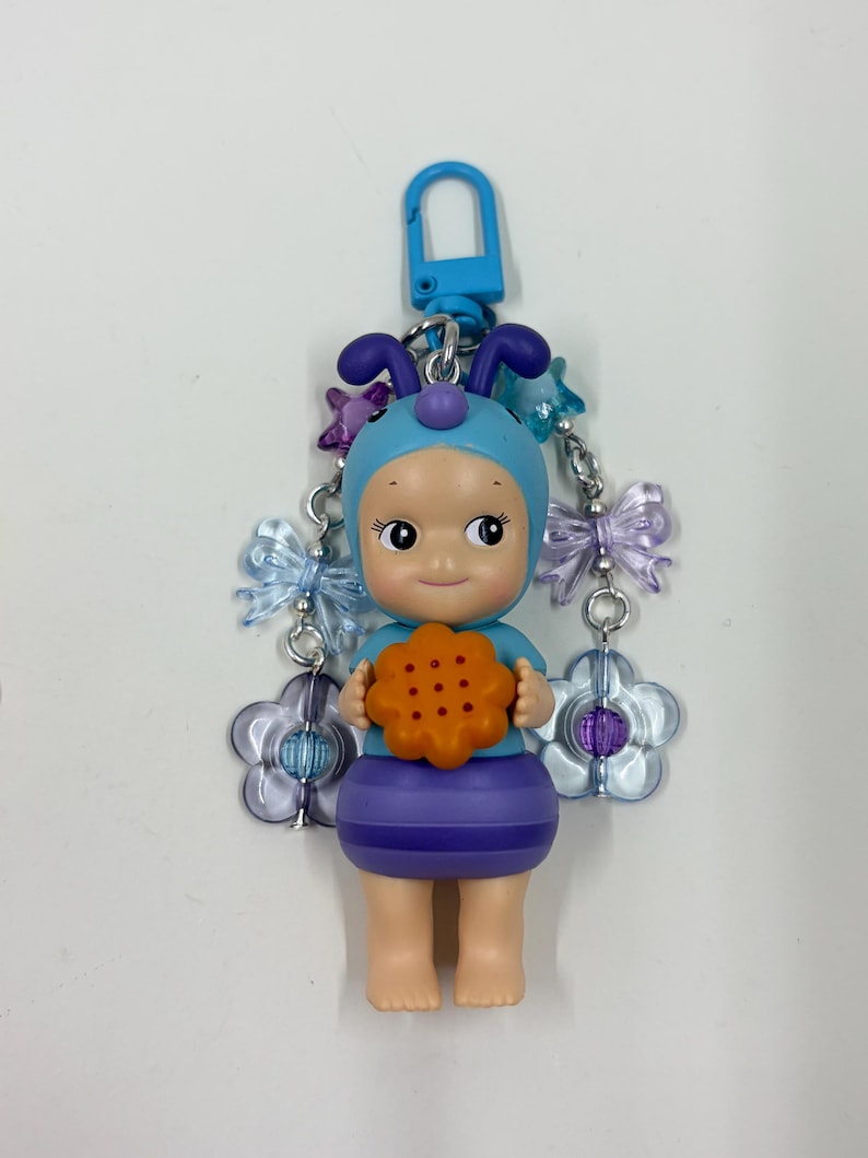 May include: A blue and purple keychain featuring a doll-like figure wearing a blue bonnet with antennae and holding an orange cookie. The keychain has a blue clasp and is adorned with flower and butterfly charms.