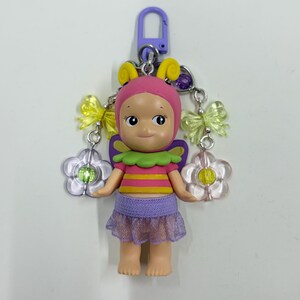 May include: A colorful keychain featuring a doll-like figure with a pink cap, yellow antennae, and a purple skirt. The figure has a green and yellow striped top, and is adorned with yellow bows and flower charms. The keychain is attached to a purple clasp.