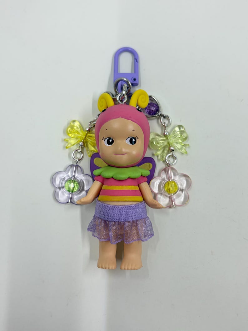 May include: A colorful keychain featuring a doll-like figure with a pink cap, yellow antennae, and a purple skirt. The figure has a green and yellow striped top, and is adorned with yellow bows and flower charms. The keychain is attached to a purple clasp.