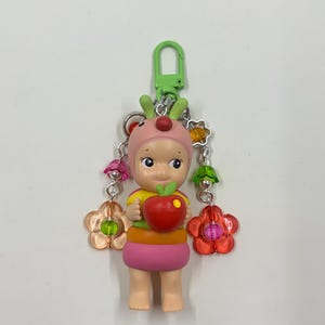 May include: A colorful keychain featuring a small figurine wearing a pink hat with red accents and holding a red apple. The figurine has a yellow and orange top and a pink skirt. The keychain includes a green clasp and decorative flower charms.