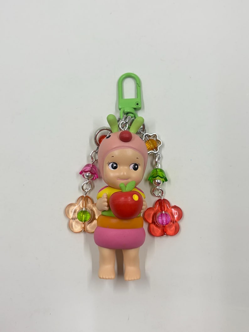 May include: A colorful keychain featuring a small figurine wearing a pink hat with red accents and holding a red apple. The figurine has a yellow and orange top and a pink skirt. The keychain includes a green clasp and decorative flower charms.