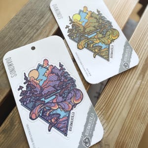 Könnte beinhalten: Zwei weiße Aufkleberbögen mit dem Wort "DIAMONDS" oben gedruckt. Jeder Bogen zeigt ein farbenfrohes, abstraktes Design mit einer Bergkette und dem Text "Riverwalker.ca" unten gedruckt. Der Text "Für beste Ergebnisse auf saubere, trockene Oberflächen auftragen" ist unten auf jedem Bogen gedruckt.
