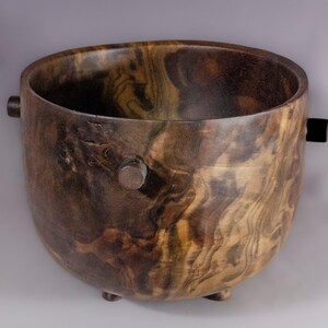 May include: A handcrafted wooden bowl with a dark brown and tan wood grain pattern. The bowl has a rounded shape with two small handles and three small feet. The interior is dark, and the exterior shows the natural wood grain.