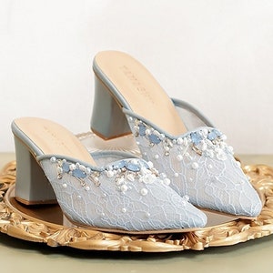 Puede incluir: Un par de mules de tacón azul claro con un delicado diseño de encaje y adornos de perlas y cristales. Los zapatos están sobre una bandeja decorativa dorada.