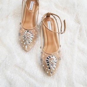 Puede incluir: Un par de zapatos planos de novia beige con correas de tobillo. Los zapatos est&aacute;n adornados con un gran grupo de diamantes de imitaci&oacute;n y perlas en la punta.