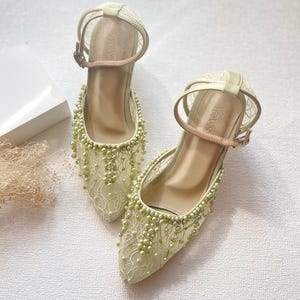 May include: A pair of light green, heeled shoes with ankle straps. The shoes are adorned with delicate lace and small, light green beads. The straps have a buckle closure. The heels are approximately 2 inches high.