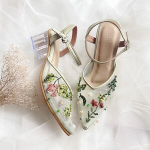 K&ouml;nnte beinhalten: Ein Paar hellgr&uuml;ne High Heels mit Blumenstickerei und durchsichtigen Abs&auml;tzen. Die Schuhe haben ein zartes Blumenmuster mit rosa, gr&uuml;nen und wei&szlig;en Akzenten. Der Markenname "YevS" ist auf dem rechten Schuh sichtbar.