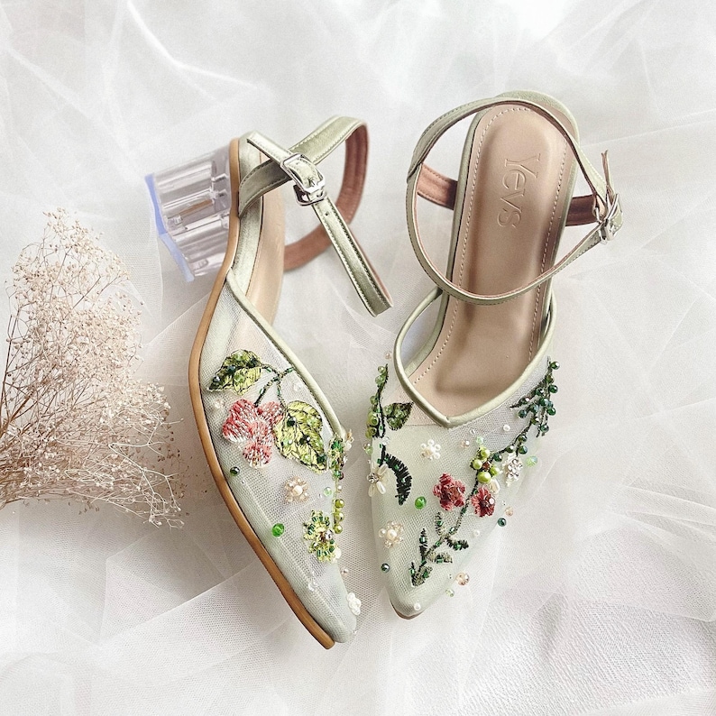 K&ouml;nnte beinhalten: Ein Paar hellgr&uuml;ne High Heels mit Blumenstickerei und durchsichtigen Abs&auml;tzen. Die Schuhe haben ein zartes Blumenmuster mit rosa, gr&uuml;nen und wei&szlig;en Akzenten. Der Markenname "YevS" ist auf dem rechten Schuh sichtbar.