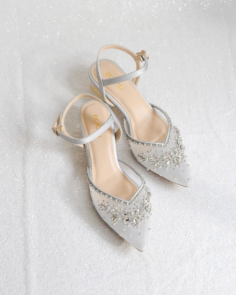 May include: A pair of silver satin bridal shoes with a pointed toe and ankle strap. The shoes are embellished with rhinestones and beads.