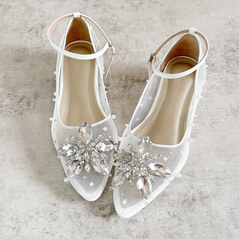 May include: A pair of white pointed-toe flats featuring a sheer mesh upper adorned with scattered small white pearls. Each shoe has a prominent floral-shaped embellishment made of clear faceted crystals at the toe and a thin ankle strap with a buckle.