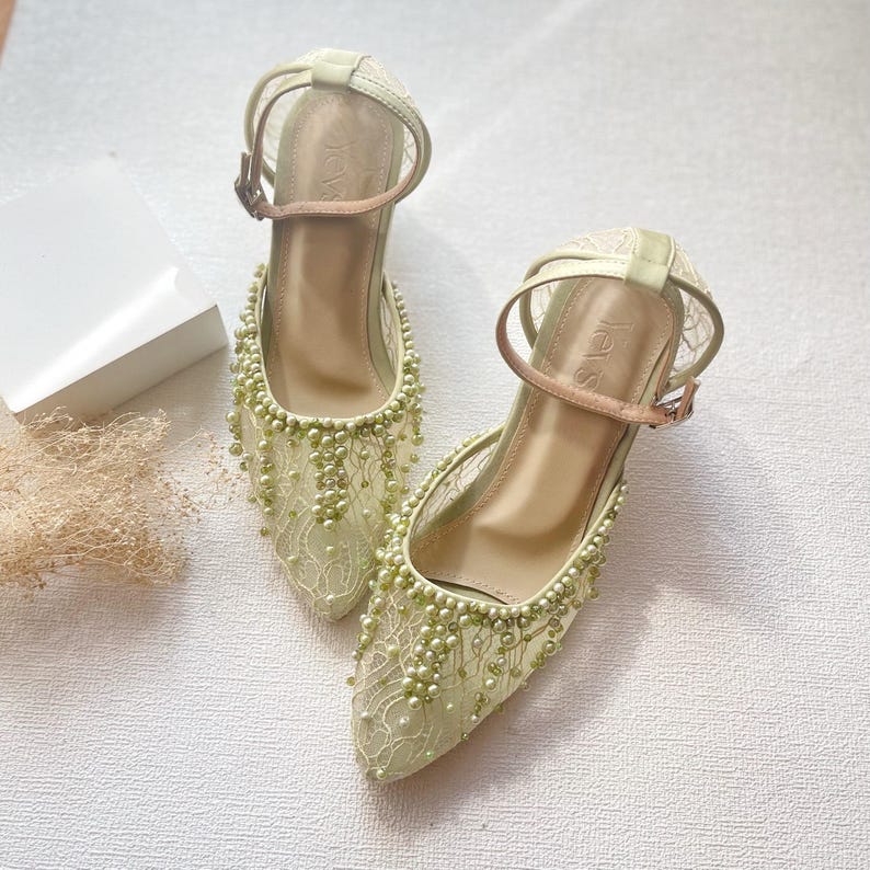 May include: A pair of light green, heeled shoes with ankle straps. The shoes are adorned with delicate lace and small, light green beads. The straps have a buckle closure. The heels are approximately 2 inches high.
