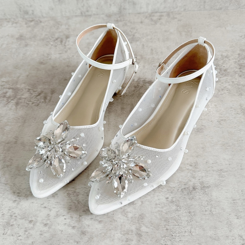 May include: White bridal shoes with a delicate mesh fabric and a large crystal flower embellishment. The shoes have a pointed toe and an ankle strap.