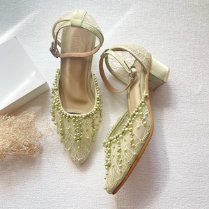 May include: A pair of light green heeled shoes with an ankle strap and a pointed toe. The shoes are adorned with delicate lace and small, dangling beads. The heels are a block style, and the straps are a light brown color.