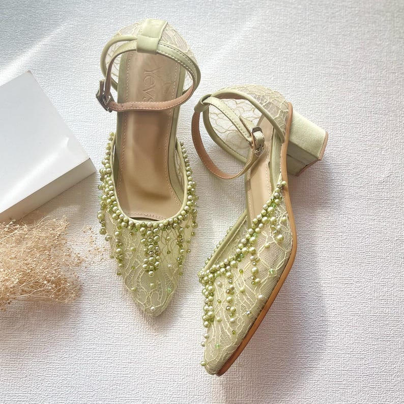 May include: A pair of light green heeled shoes with an ankle strap and a pointed toe. The shoes are adorned with delicate lace and small, dangling beads. The heels are a block style, and the straps are a light brown color.