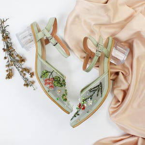 K&ouml;nnte beinhalten: Ein Paar hellgr&uuml;ne Sandalen mit hohen Abs&auml;tzen und transparenten Acryl-Abs&auml;tzen. Die Sandalen sind mit floralen Stickereien und Perlen verziert.