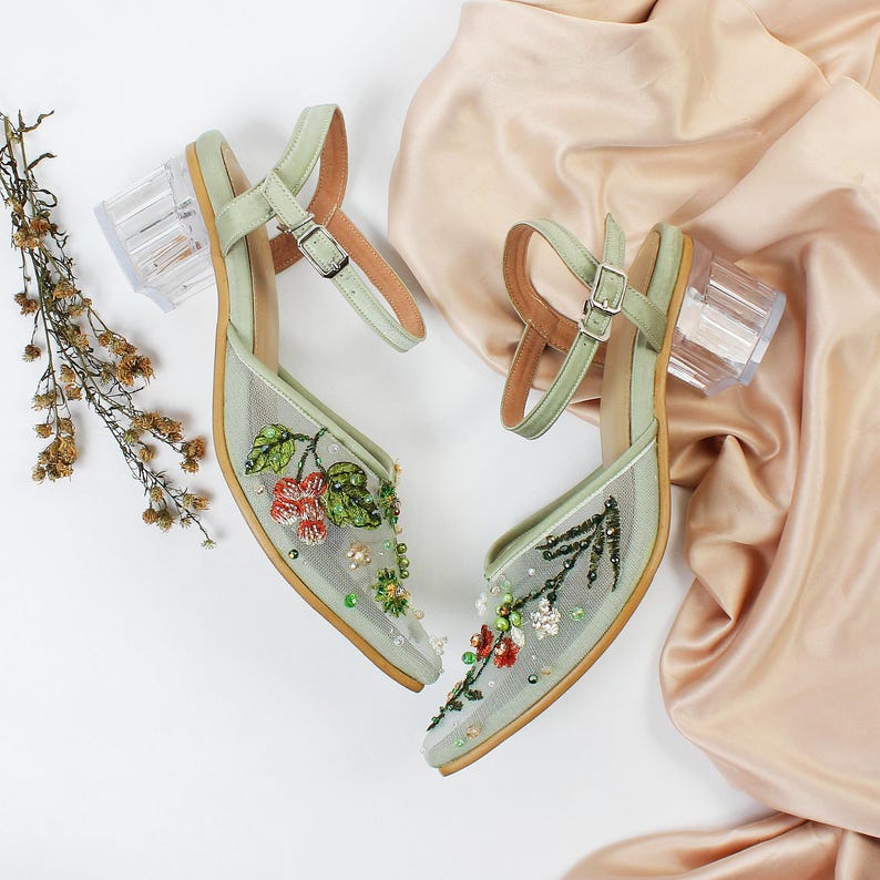 K&ouml;nnte beinhalten: Ein Paar hellgr&uuml;ne Sandalen mit hohen Abs&auml;tzen und transparenten Acryl-Abs&auml;tzen. Die Sandalen sind mit floralen Stickereien und Perlen verziert.