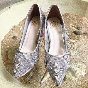 May include: A pair of silver high-heeled shoes with a lace and rhinestone design. The shoes have a pointed toe and a delicate heel. The brand name "FLAVA" is visible on the inside of the shoes.