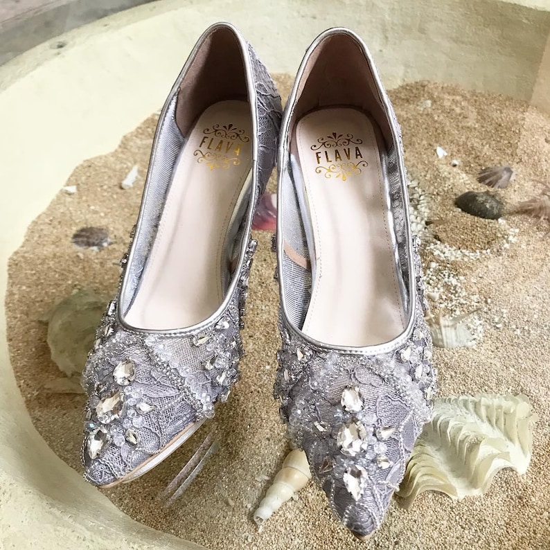 May include: A pair of silver high-heeled shoes with a lace and rhinestone design. The shoes have a pointed toe and a delicate heel. The brand name "FLAVA" is visible on the inside of the shoes.