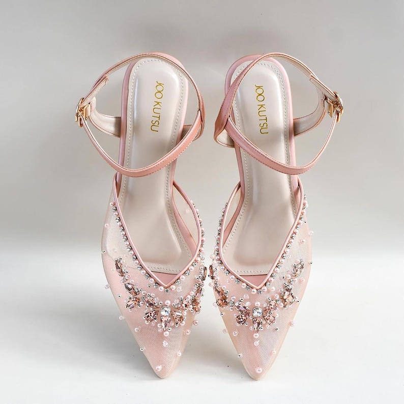 May include: A pair of blush pink high-heeled shoes with delicate mesh fabric and embellished with sparkling rhinestones and pearls. The shoes have ankle straps with gold buckles and the brand name "JOO KUTSU" is printed on the inside of the straps.