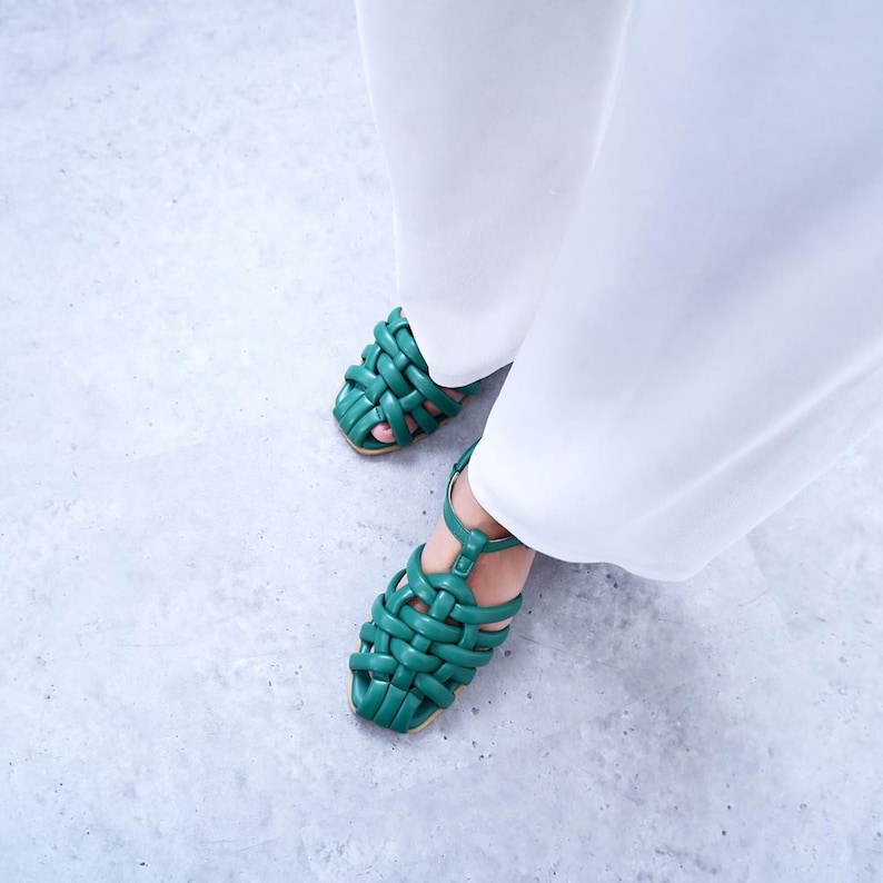 May include: A pair of green woven leather sandals with ankle straps. The sandals are being worn by someone wearing white trousers.