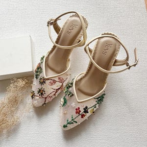 Puede incluir: Sandalias beige bordadas con diseños florales en rosa, verde y amarillo. Las sandalias tienen punta afilada, correas en el tobillo y la marca "Yevs" en la plantilla.