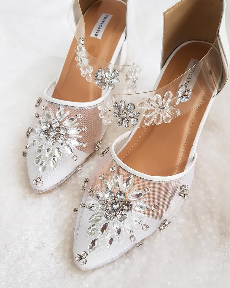 Puede incluir: Zapatos de novia blancos con punta puntiaguda y parte superior de malla. Los zapatos est&aacute;n decorados con grandes diamantes de imitaci&oacute;n transparentes y tienen una correa transparente con la palabra "UNICATIE" escrita en ella.
