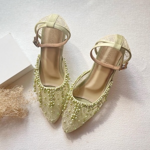 May include: A pair of light green, pointed-toe heels with ankle straps. The shoes feature a delicate lace overlay and are embellished with small, light green beads. The straps are a light tan color, and the heels have the word "Yevs" printed on them.