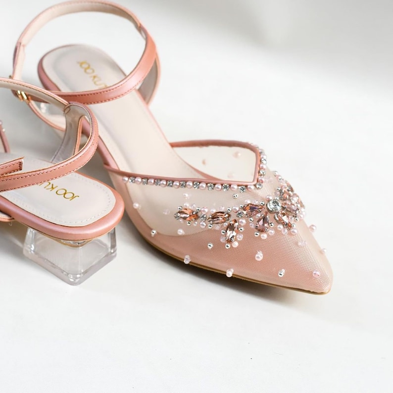 May include: A pair of blush pink mesh heeled shoes with a delicate floral design made of rhinestones and pearls. The shoes have a clear acrylic heel.