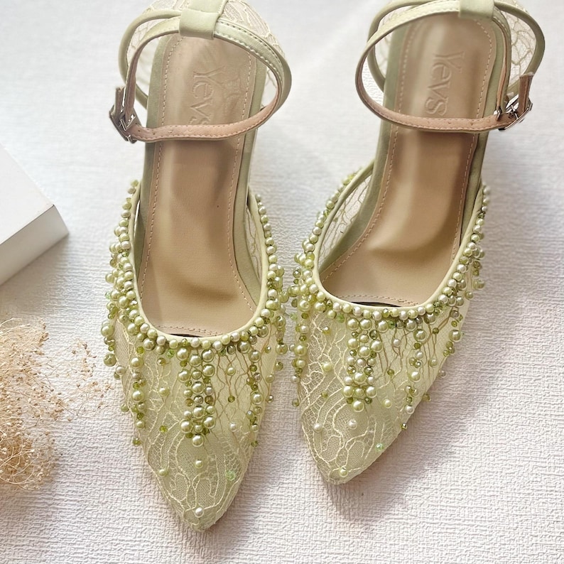 May include: A pair of pale green pointed-toe heels with ankle straps. The shoes feature a delicate lace overlay adorned with small, light green beads. The heels have a medium height and a buckle closure.