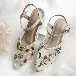 K&ouml;nnte beinhalten: Ein Paar hellgr&uuml;ne High-Heel-Schuhe mit zarter Blumenstickerei und einem Mesh-Gewebe. Die Schuhe haben eine spitze Zehenpartie und einen Kn&ouml;chelriemen mit einer Schnalle. Der Markenname "Yev's" ist auf der Innenseite der Schuhe sichtbar.
